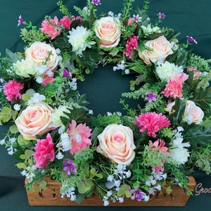 May include: A floral wreath featuring peach roses, pink and white chrysanthemums, and small purple flowers. The wreath is set on a wooden base with the text "GroovyRuby" on the bottom right. The wreath is surrounded by green foliage.