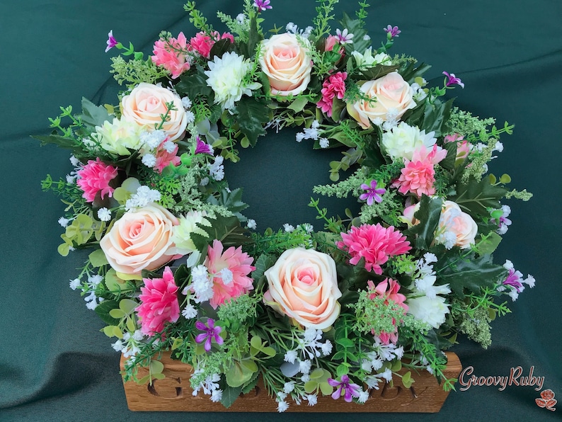 May include: A floral wreath featuring peach roses, pink and white chrysanthemums, and small purple flowers. The wreath is set on a wooden base with the text "GroovyRuby" on the bottom right. The wreath is surrounded by green foliage.