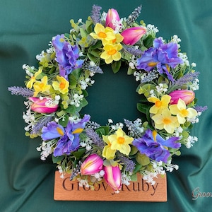 May include: A colourful floral wreath with purple irises, yellow daffodils, and pink tulips. The wreath is decorated with lavender and small white flowers. A wooden sign at the base reads "Groovy Ruby".