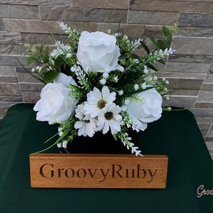 May include: A white artificial flower arrangement with roses and daisies in a black vase. The arrangement is on a green surface with a wooden sign that reads "GroovyRuby".