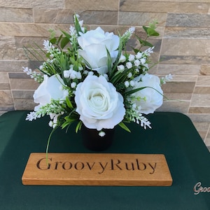 May include: A white artificial flower arrangement in a black pot, with a wooden sign that reads "GroovyRuby".