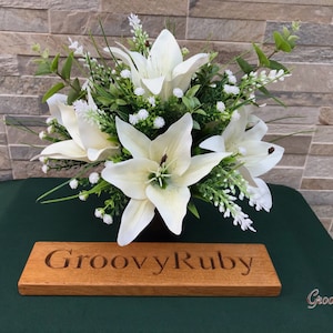 May include: A white artificial lily arrangement in a black vase with a wooden sign that reads "GroovyRuby".