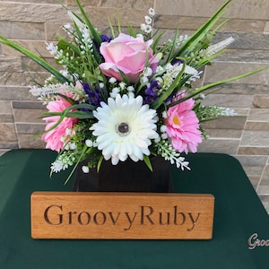 May include: An artificial flower arrangement in a black box with a wooden sign that reads "GroovyRuby". The arrangement features pink, white, and green flowers.