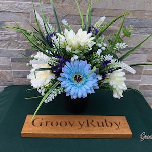 May include: A blue and white artificial flower arrangement in a black vase. The arrangement is on a green table with a wooden sign that reads "Groovy Ruby".