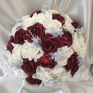 May include: A bridal bouquet featuring ivory and burgundy roses, accented with silver glitter and sprigs. The bouquet is arranged in a round shape and features a variety of textures.