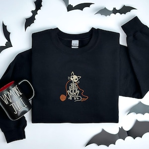 Könnte beinhalten: Ein schwarzes Langarm-Sweatshirt mit einer Stickerei eines weißen Katzenskeletts, das mit einem orangefarbenen Wollknäuel spielt. Neben dem Sweatshirt steht eine schwarze Tasse mit rotem Innenraum und dem weißen Text VAMPIRE JUICE.