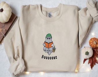 May include: A beige sweatshirt with a ghost wearing a green hat reading a book and holding a cup of coffee. The text "BOO-OOOKS" is printed below the ghost.