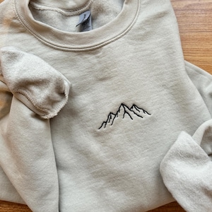 Könnte beinhalten: Ein hellbeige Sweatshirt mit Rundhalsausschnitt und langen Ärmeln. Auf der Brust befindet sich ein kleiner, gestickter Bergkamm aus schwarzem Faden. Der Stoff wirkt weich und bequem, geeignet für Freizeitkleidung.