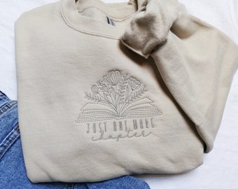 May include: A beige sweatshirt with an embroidered design of an open book with flowers growing out of it. The text "Just one more chapter" is embroidered below the book.