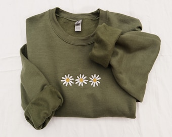 Könnte beinhalten: Ein grüner Pullover mit drei gestickten weißen Gänseblümchen mit gelben Zentren.
