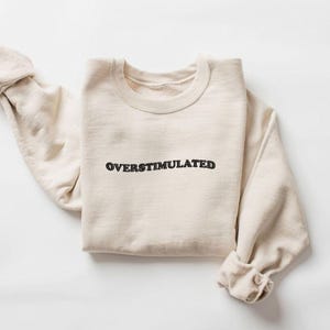 May include: A cream-colored sweatshirt with the word "OVERSTIMULATED" embroidered in black across the chest. The sleeves are rolled up, and the garment is laid flat on a white surface. The sweatshirt has a crew neck.