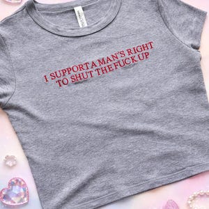 Könnte beinhalten: Ein graues, meliertes bauchfreies T-Shirt mit dem roten Schriftzug I SUPPORT A MAN'S RIGHT TO SHUT THE FUCK UP auf der Brust. Das Kleidungsstück hat einen Rundhalsausschnitt und kurze Ärmel und besticht durch ein schlichtes Design.