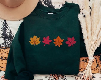 Puede incluir: Una sudadera verde oscuro con una fila de cuatro hojas de arce de colores impresas en la parte delantera. Las hojas son de color naranja, rojo y amarillo.