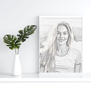 May include: A black and white pencil drawing of a woman with long hair smiling. The drawing is framed in a white frame and is hanging on a white wall.