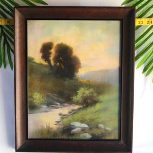 Puede incluir: Pintura de paisaje enmarcada que representa una escena serena con un arroyo, árboles y colinas. La obra de arte presenta una paleta de colores cálidos con tonos de verde, marrón y amarillo. El marco es de color marrón oscuro. La pintura mide aproximadamente 40 cm por 61 cm.
