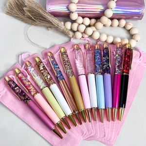 May include: A set of 12 glitter pens with gold accents and clear barrels filled with colorful glitter. The pens are arranged in a fan shape and are displayed on a pink velvet pouch.