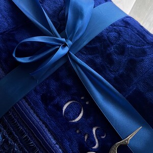 Peut inclure: Une couverture en peluche bleu roi pli&eacute;e avec un motif floral en relief, nou&eacute;e par un ruban en satin assorti. La couverture pr&eacute;sente une bordure &agrave; franges, les lettres O et S brod&eacute;es, et une paire de ciseaux argent&eacute;s en forme de cigogne.