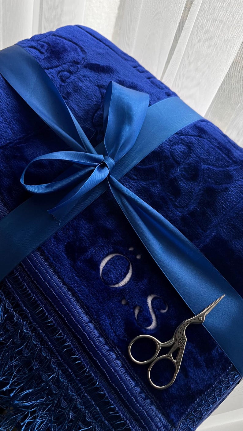 Peut inclure: Une couverture en peluche bleu roi pli&eacute;e avec un motif floral en relief, nou&eacute;e par un ruban en satin assorti. La couverture pr&eacute;sente une bordure &agrave; franges, les lettres O et S brod&eacute;es, et une paire de ciseaux argent&eacute;s en forme de cigogne.