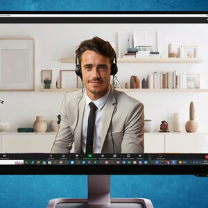 May include: A man wearing a beige suit jacket and a black tie is on a video call. He is wearing a headset and smiling at the camera. The background is a white wall with a bookshelf and a potted plant.