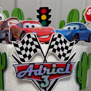 May include: A white cake decorated with a red race car, a blue race car, a chequered flag, a traffic light, a stop sign, and a green cactus. The cake is topped with a red and silver shield with the name "Adriel" and the number "3" on it.