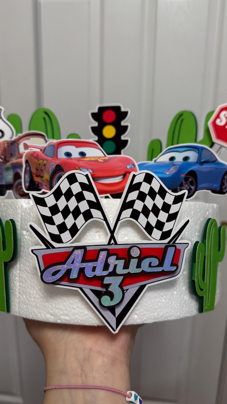 May include: A white cake decorated with a red race car, a blue race car, a chequered flag, a traffic light, a stop sign, and a green cactus. The cake is topped with a red and silver shield with the name "Adriel" and the number "3" on it.
