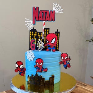 Peut inclure: Un gâteau bleu givré décoré de découpes sur le thème de Spider-Man. Le gâteau a une base dorée et présente le nom "Natan" et le chiffre "6". Des figurines de Spider-Man sont placées autour du gâteau.
