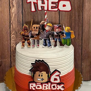 Może przedstawiać: Biało-czerwony tort z motywem Roblox z postacią Roblox z przodu. Tort jest ozdobiony czerwono-białym patyczkiem, czerwono-białym napisem "THEO" i kilkoma postaciami Roblox. Tort stoi na złotej podstawce.