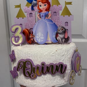 May include: A purple and gold cake topper featuring a princess from the cartoon 'Sofia the First'. The topper includes a castle, a bird, a squirrel, a rabbit, and the number 3. The cake is decorated with a purple and gold 'Quinn' name topper and a carriage.