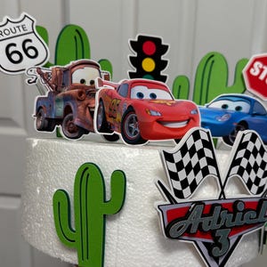May include: A white cake decorated with a Cars theme. The cake is decorated with a red race car, a tow truck, a stop sign, a traffic light, a cactus, and a chequered flag. The cake also has a sign that says "Route 66" and a sign that says "Adriel 3".