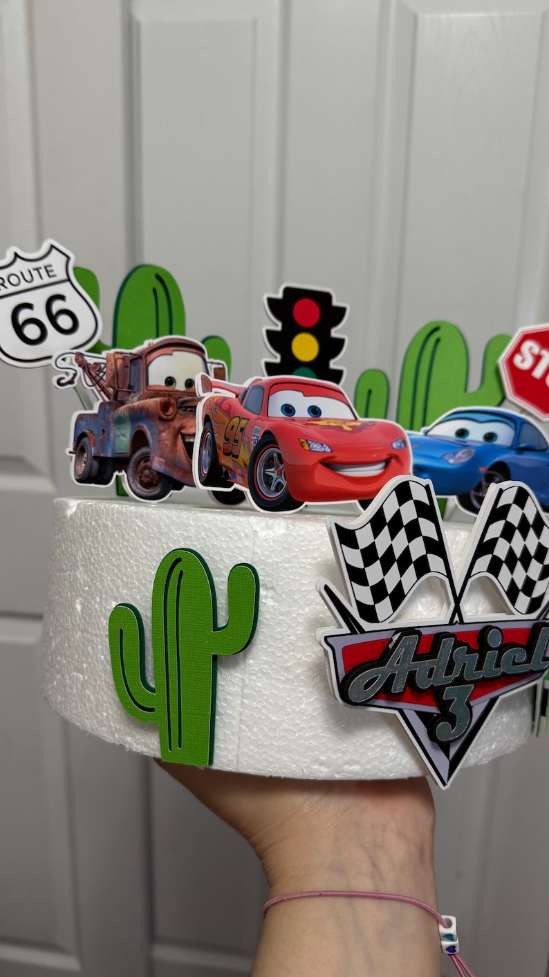May include: A white cake decorated with a Cars theme. The cake is decorated with a red race car, a tow truck, a stop sign, a traffic light, a cactus, and a chequered flag. The cake also has a sign that says "Route 66" and a sign that says "Adriel 3".