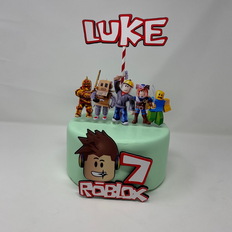 Może przedstawiać: Zielony tort z ozdobą w stylu Roblox. Ozdoba zawiera czerwony i biały pasek w paski z czerwonym banerem z napisem "LUKE" oraz zieloną ozdobę tortu z postacią Roblox i napisem "ROBLOX".