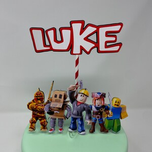 Może przedstawiać: Topper na tort z napisem "LUKE" w czerwonych i białych literach z czerwono-białym paskiem. Topper zawiera r&oacute;wnież pięć postaci z Roblox: rycerza, robota, robotnika budowlanego, pirata i postać w niebieskiej koszuli.