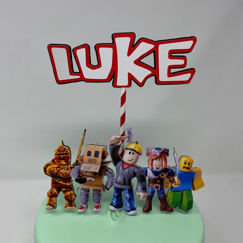 Może przedstawiać: Topper na tort z napisem "LUKE" w czerwonych i białych literach z czerwono-białym paskiem. Topper zawiera r&oacute;wnież pięć postaci z Roblox: rycerza, robota, robotnika budowlanego, pirata i postać w niebieskiej koszuli.