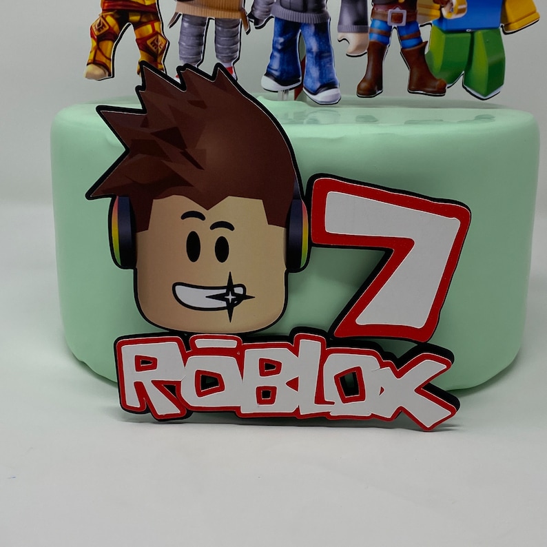 Może przedstawiać: Zielony tort z czerwono-białym logo Roblox, czerwoną cyfrą 7 i kresk&oacute;wkową postacią Roblox z brązowymi włosami i słuchawkami.
