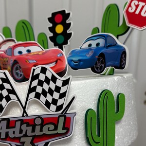 May include: A white cake decorated with a red car, a blue car, a stop sign, a traffic light, a chequered flag, a cactus, and the name "Adriel 3".