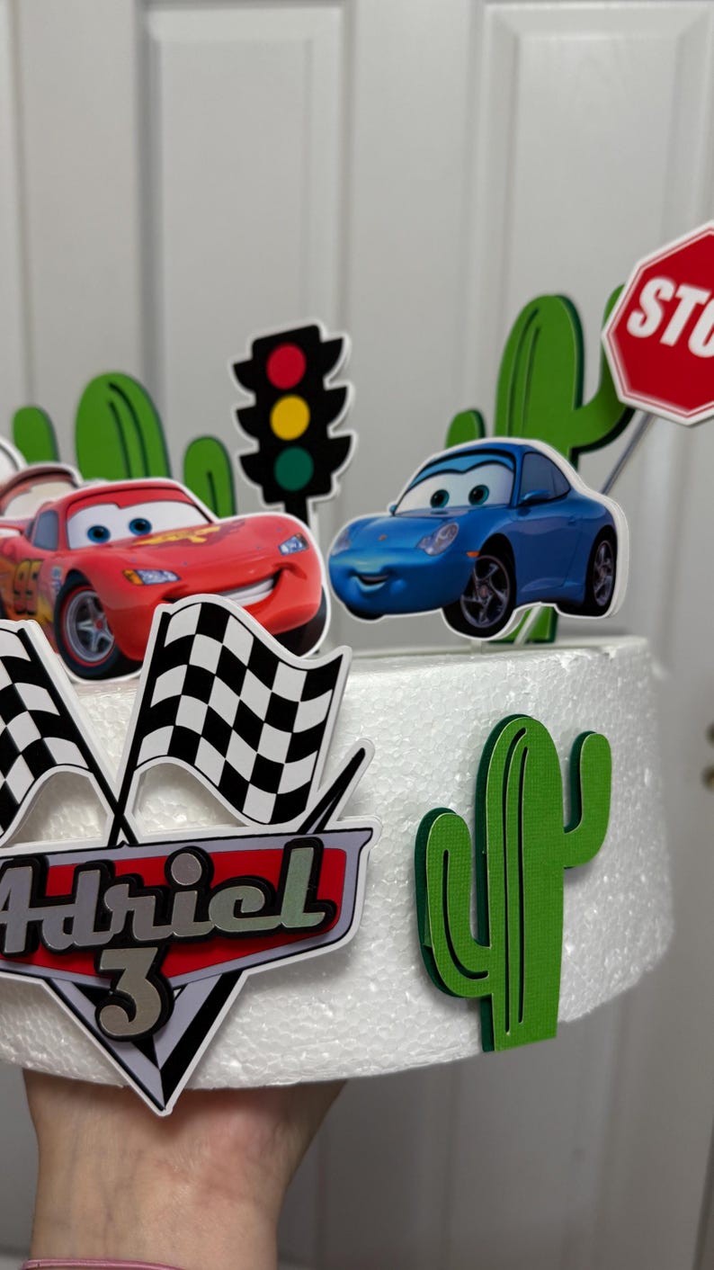 May include: A white cake decorated with a red car, a blue car, a stop sign, a traffic light, a chequered flag, a cactus, and the name "Adriel 3".