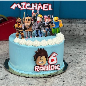 Może przedstawiać: Niebieski i biały tort urodzinowy z motywem Roblox. Tort jest ozdobiony postacią Roblox, słowem "Roblox" i liczbą "6". Tort ma r&oacute;wnież topper z imieniem "Michael" i kilkoma postaciami Roblox.