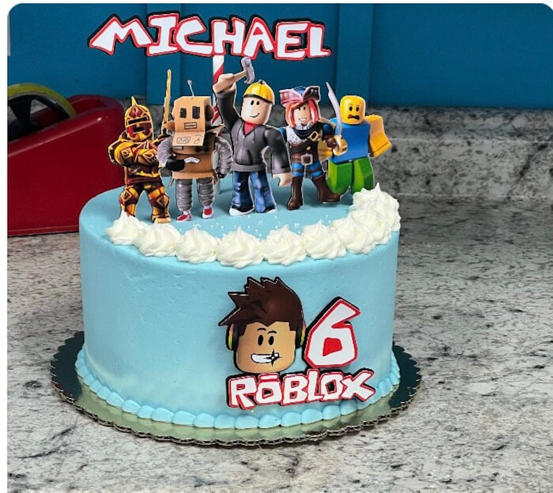 Może przedstawiać: Niebieski i biały tort urodzinowy z motywem Roblox. Tort jest ozdobiony postacią Roblox, słowem "Roblox" i liczbą "6". Tort ma r&oacute;wnież topper z imieniem "Michael" i kilkoma postaciami Roblox.