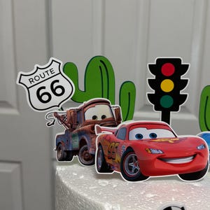 May include: A white cake decorated with a red race car, a green cactus, a red and yellow traffic light, a blue car, a chequered flag, and a sign that says "Route 66".