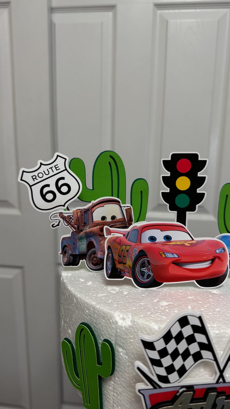 May include: A white cake decorated with a red race car, a green cactus, a red and yellow traffic light, a blue car, a chequered flag, and a sign that says "Route 66".