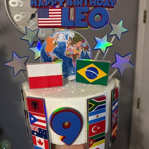 May include: A birthday cake with a blue "Happy Birthday LEO" topper, an American flag, and a globe. The cake is decorated with flags from various countries, silver star decorations, and the number 9.