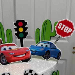 May include: A white cake decorated with a red car, a blue car, a stop sign, a traffic light, and green cacti. The cake is also decorated with a chequered flag and the name "Adriel".