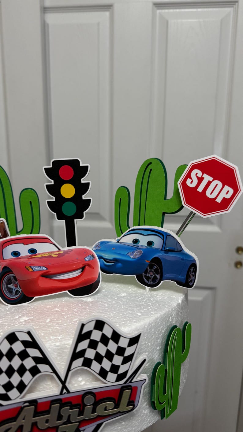 May include: A white cake decorated with a red car, a blue car, a stop sign, a traffic light, and green cacti. The cake is also decorated with a chequered flag and the name "Adriel".