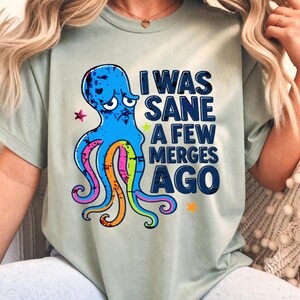 Puede incluir: Una camiseta de color verde salvia claro con un gráfico de un pulpo azul angustiado con tentáculos coloridos. El texto impreso en azul oscuro dice: I WAS SANE A FEW MERGES AGO. El diseño incluye pequeños detalles de estrellas rosas, amarillas y naranjas.