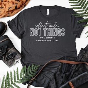 May include: A dark gray short-sleeve t-shirt featuring the white text "collect miles NOT THINGS" followed by "TWO WHEELS ENDLESS HORIZONS" in a clean, sans-serif font. The shirt has a crew neckline and is made of a solid, opaque fabric.