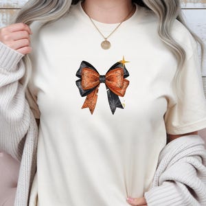 May include: A cream-coloured short-sleeve t-shirt featuring a central graphic of a decorative bow. The bow has alternating black and orange sections, each detailed with a fine white spiderweb pattern, and is accented by a small yellow star icon.