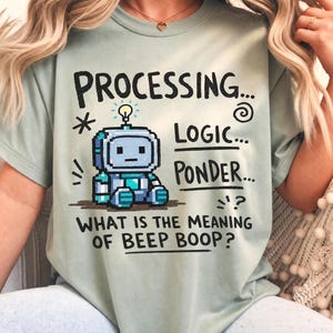 May include: A sage green t-shirt featuring a pixelated blue robot graphic with a lightbulb above its head. The text reads: PROCESSING... LOGIC... PONDER... WHAT IS THE MEANING OF BEEP BOOP? The design includes small decorative symbols like stars and swirls.