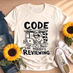以下が含まれることがあります： クリーム色の半袖Tシャツ。ヘッドランプを装着したアライグマが机の下でノートパソコンを操作している黒いグラフィックが描かれています。テキストは「CODE LIKE NOBODY'S REVIEWING」とあり、周囲にはスナックやケーブルが散らばっています。