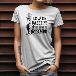 May include: A light gray t-shirt featuring a black graphic print of a cartoon character with a potted plant for a head, five small potted plants, and the text "LOW ON BASELINE DOPAMINE" in a bold, sans-serif font centered on the chest area.