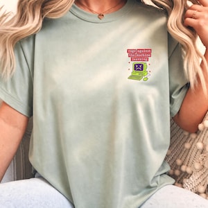 May include: A light sage green t-shirt featuring a small graphic on the upper left chest. The design shows a retro lime green computer with an angry face on the screen, accompanied by the red text "RAGE AGAINST THE MACHINE LEARNING".
